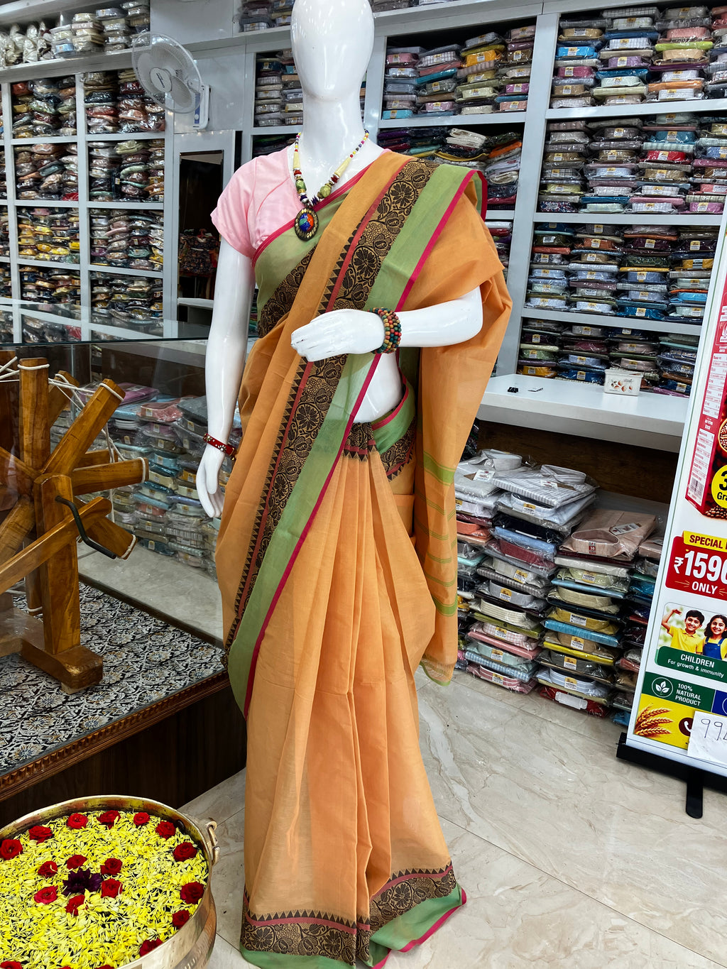 An orange saree with a green border and black patterned detailing, paired with a pink blouse, displayed on a white mannequin inside a clothing store.
