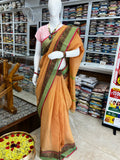 An orange saree with a green border and black patterned detailing, paired with a pink blouse, displayed on a white mannequin inside a clothing store.