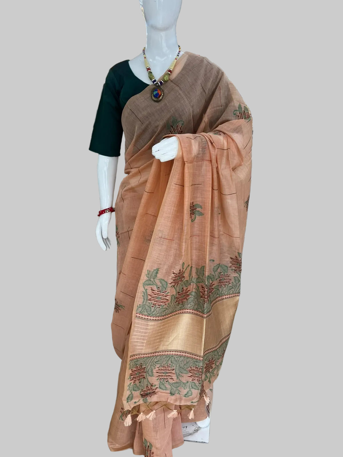 A peach-colored cotton saree with green and gold zari embroidery and border motifs, displayed on a white mannequin in a clothing store. The mannequin is also wearing a dark green blouse.