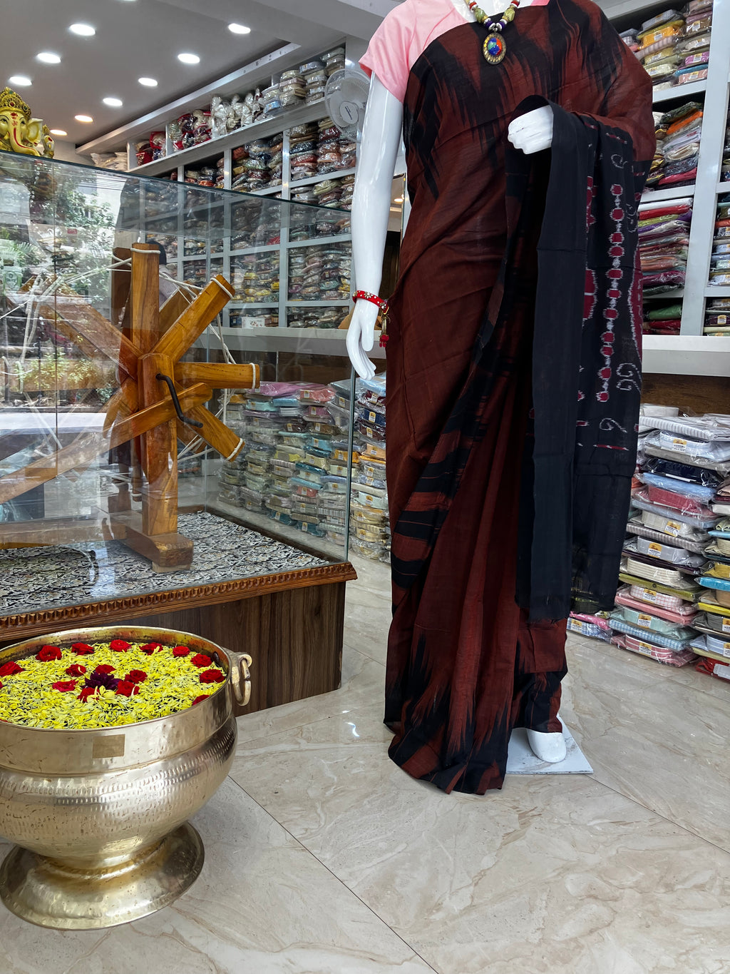 A mannequin wearing a dark brown and black sari with red and gold border details, standing inside a fabric and apparel store with shelves of folded garments in the background.