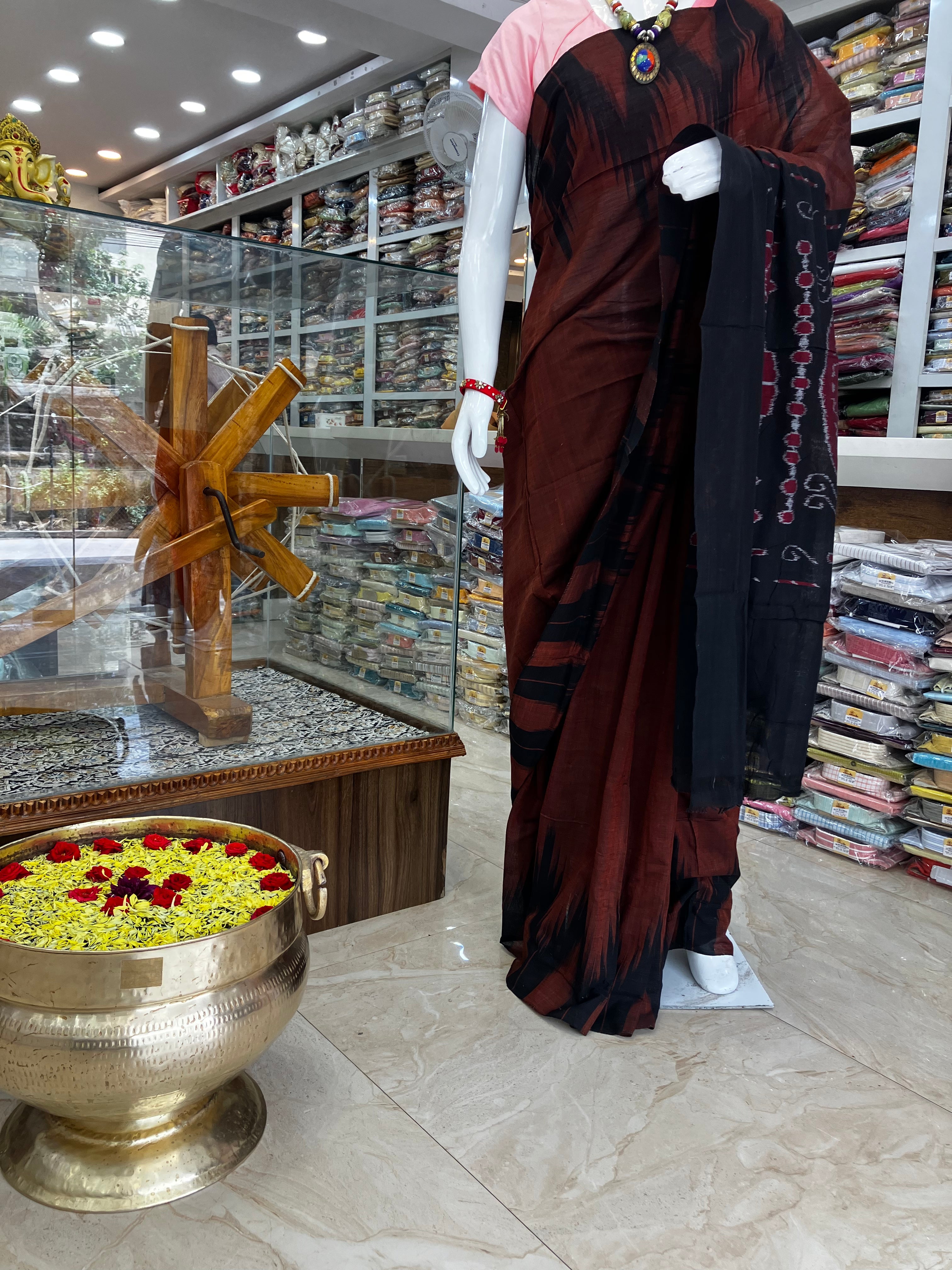 A mannequin wearing a dark brown and black sari with red and gold border details, standing inside a fabric and apparel store with shelves of folded garments in the background.