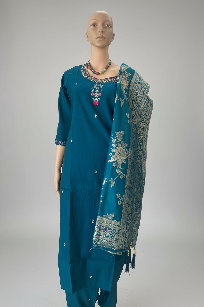 A teal blue ethnic salwar kameez suit displayed on a mannequin, featuring a heavily embroidered neckline with red and gold details, a matching printed dupatta, and small silver mirror work.